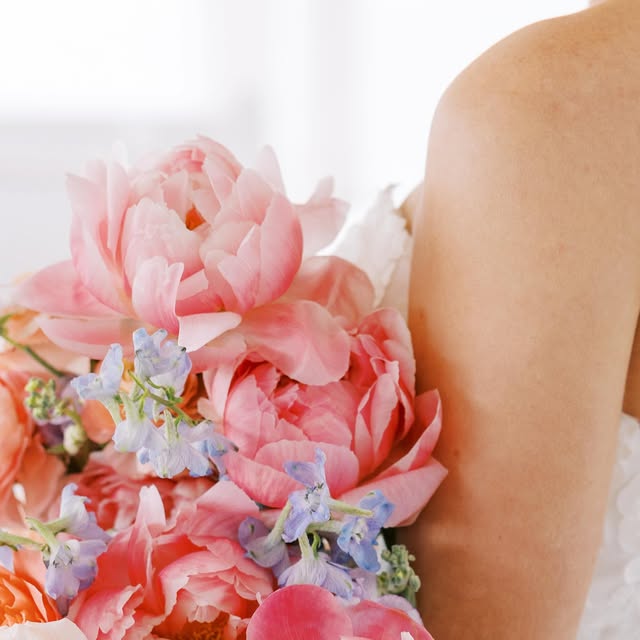 Coral peony and blue delphinium bridal bouquet resting against a bride's shoulder