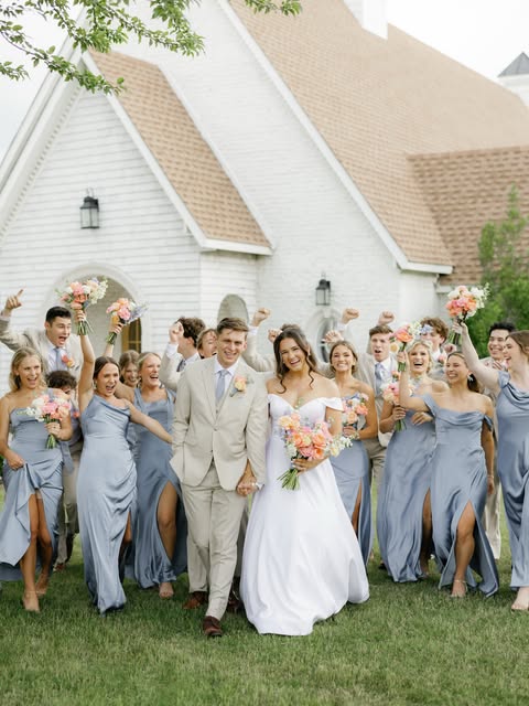 Wedding party in dusty blue celebrating with the couple outside a white chapel